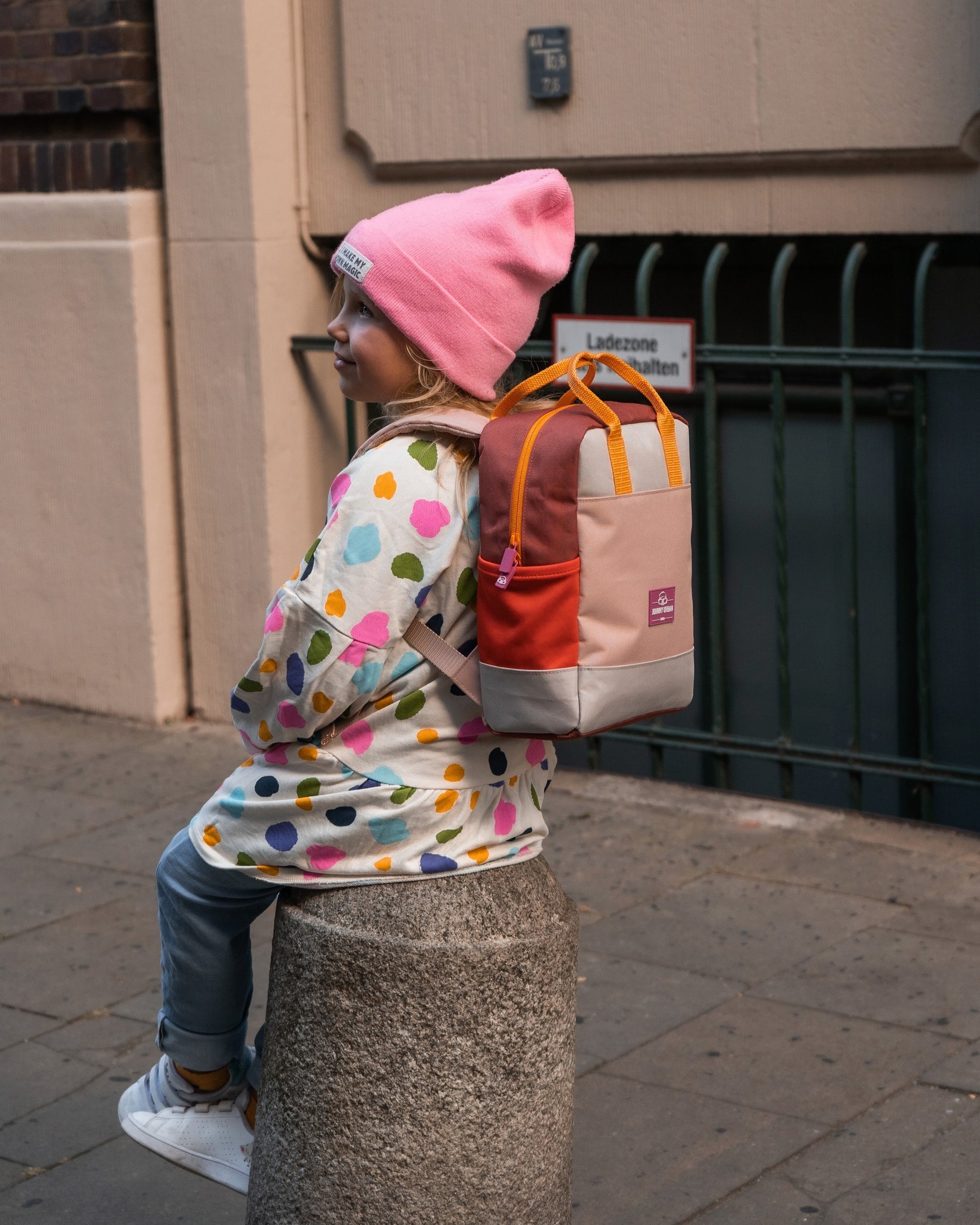 Mädchen trägt Kinderrucksack 'Linus' in Rosa mit bunten Farben, sitzt auf Mauerpfosten