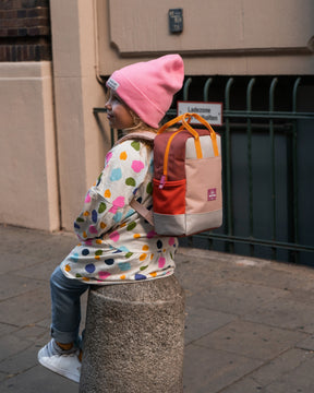 Mädchen trägt Kinderrucksack 'Linus' in Rosa mit bunten Farben, sitzt auf Mauerpfosten