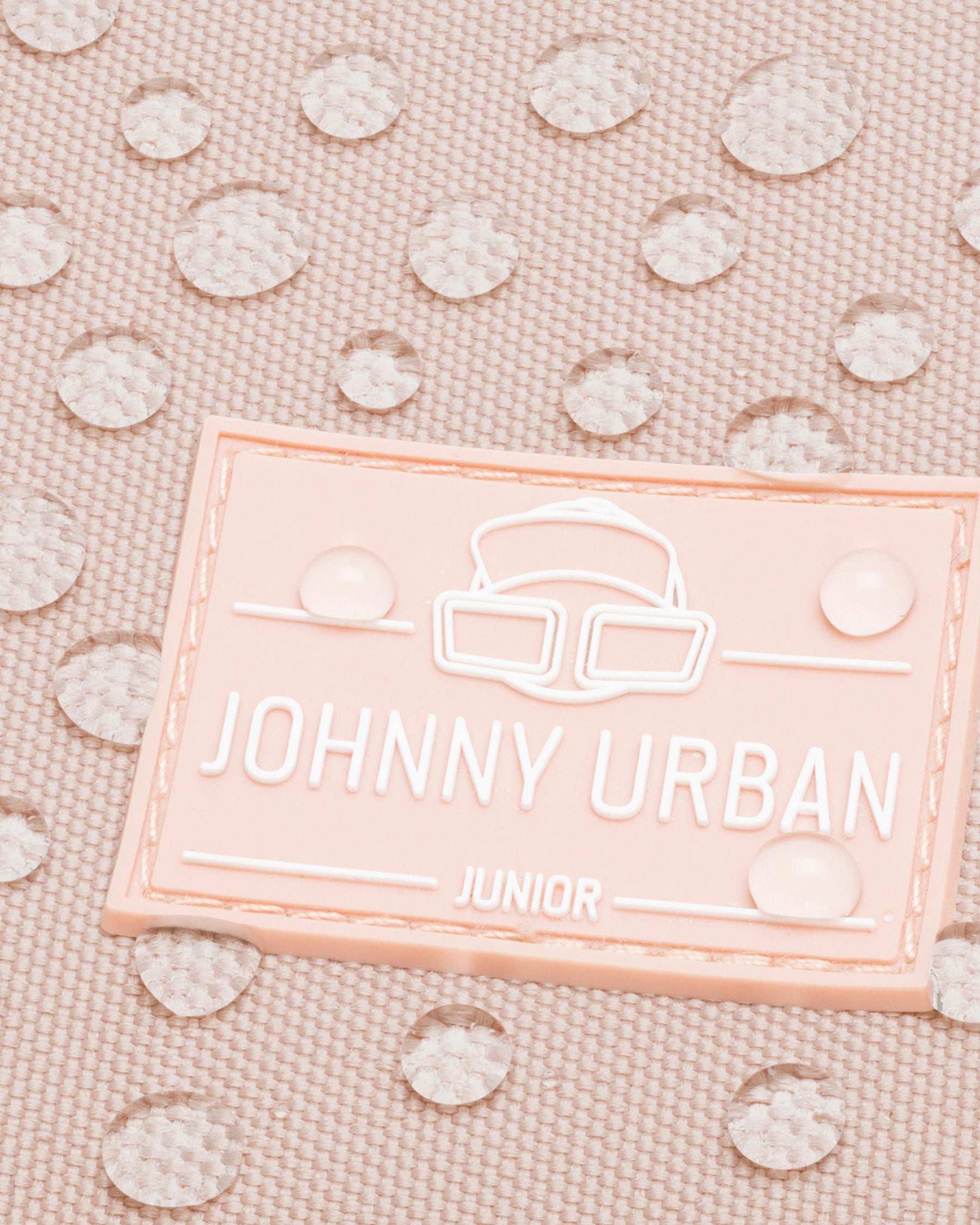 Nahaufnahme des rosa Johnny Urban Junior Logos auf wasserdichtem Stoff mit Wassertropfen