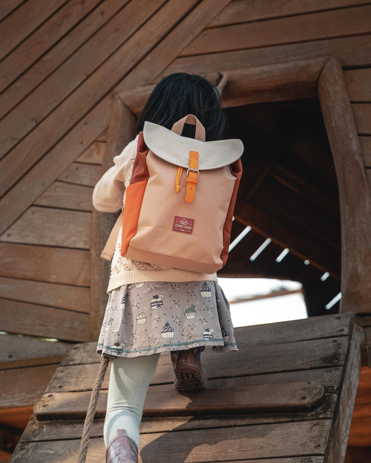 Mädchen klettert in ein Holzhaus mit rosa-buntem Kinderrucksack 'Liam' von Johnny Urban