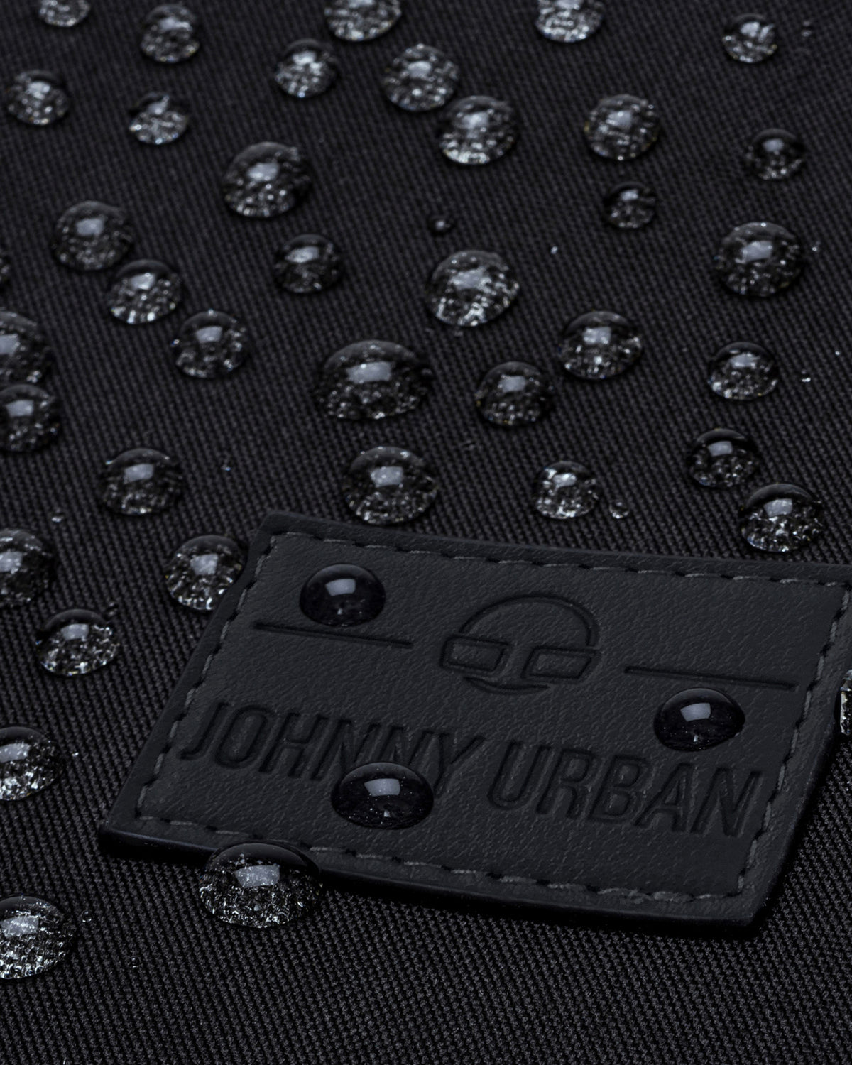 Nahe Aufnahme des schwarzen Labels des Johnny Urban Rucksacks Jona Large mit Wassertropfen auf schwarzem Stoff
