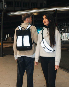Mann trägt reflektierenden schwarzen Rucksack 'Mika', Frau trägt reflektierende Umhängetasche bei Nacht in Stadt