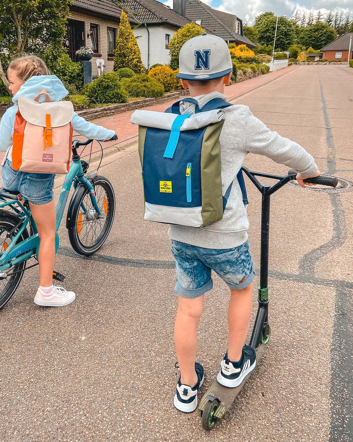 Zwei Kinder mit Rolltop-Kinderrucksäcken Aaron, ein Mädchen auf einem Fahrrad mit rosa-orangenem Rucksack, ein Junge auf einem Roller mit blau-grünem Rucksack