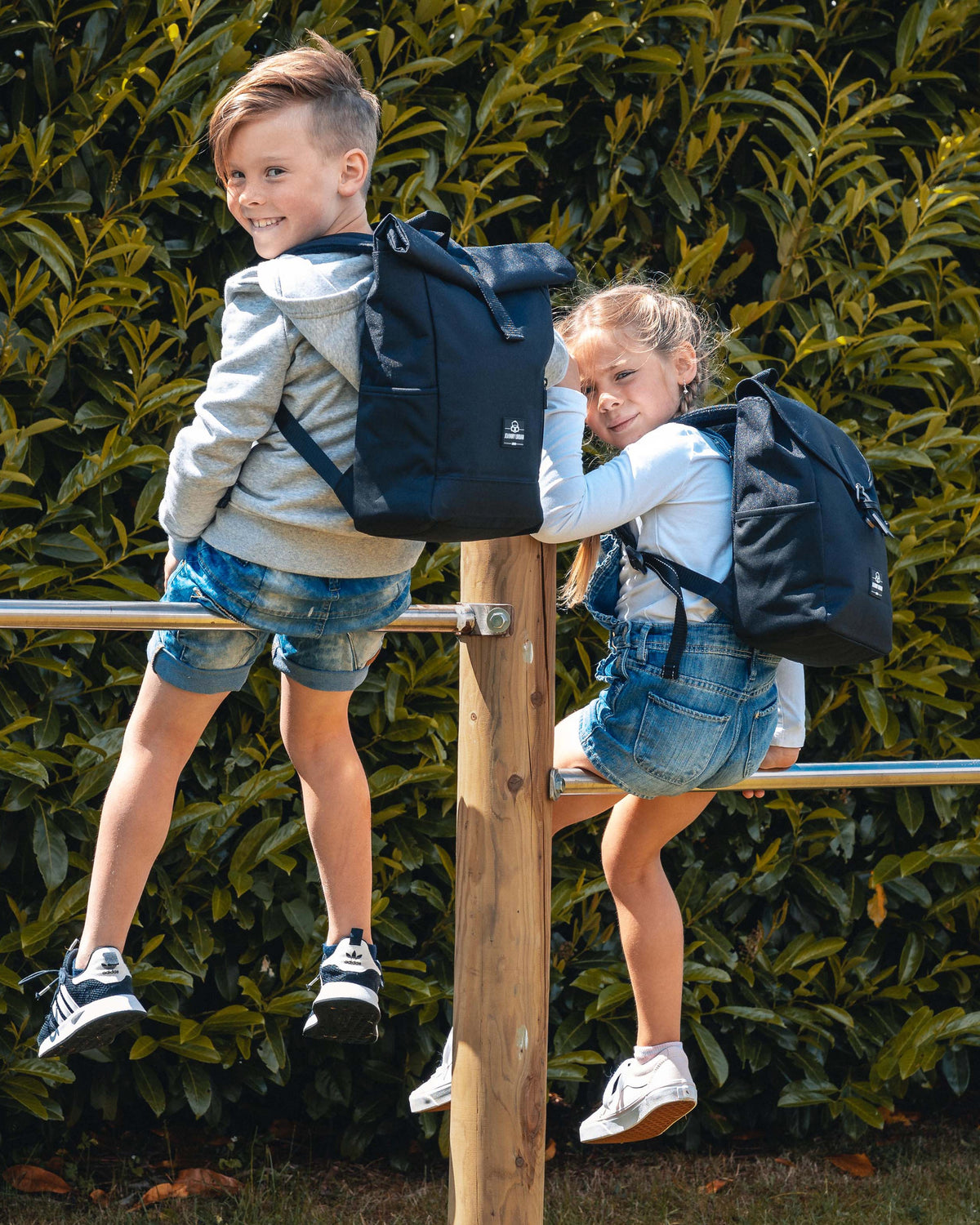 Zwei Kinder mit schwarzen Rolltop-Kinderrucksäcken ‚Aaron‘ klettern auf ein Holzgeländer im Freien