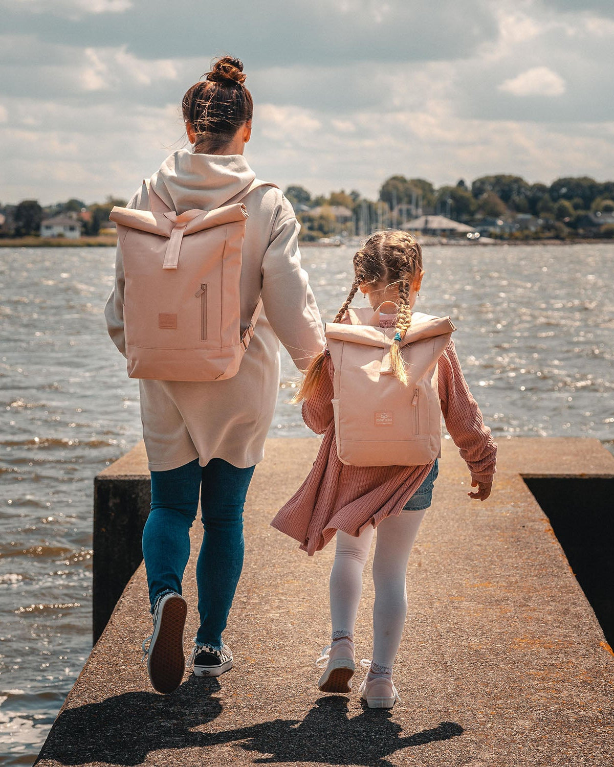 Mutter und Kind laufen auf einem Steg am Wasser, tragen rosa Rolltop Kinderrucksäcke von Johnny Urban