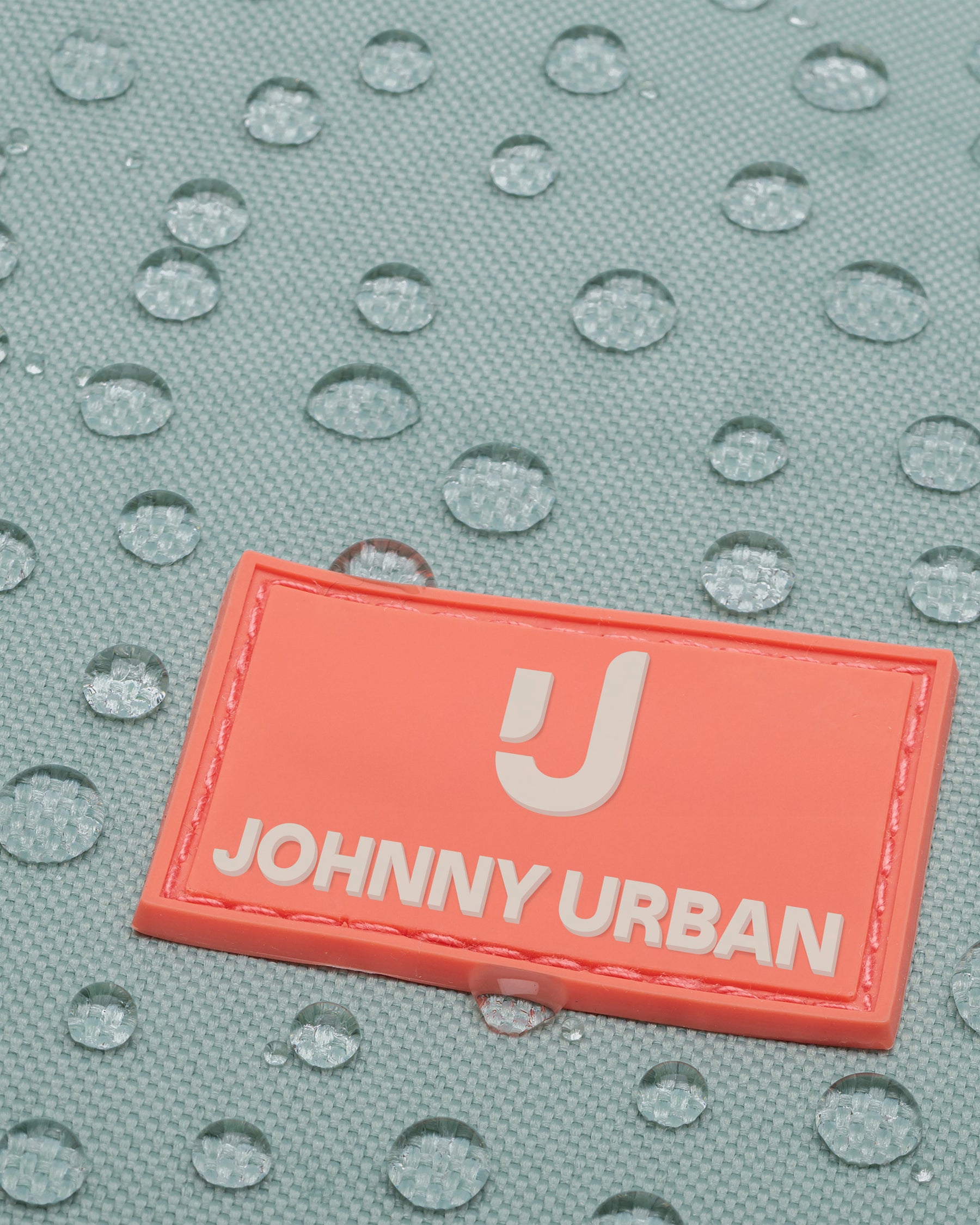 Nahaufnahme des wasserabweisenden Materials des hellblauen Rolltop Kinderrucksacks 'Aaron' mit einem orangefarbenen Johnny Urban Junior Logo.