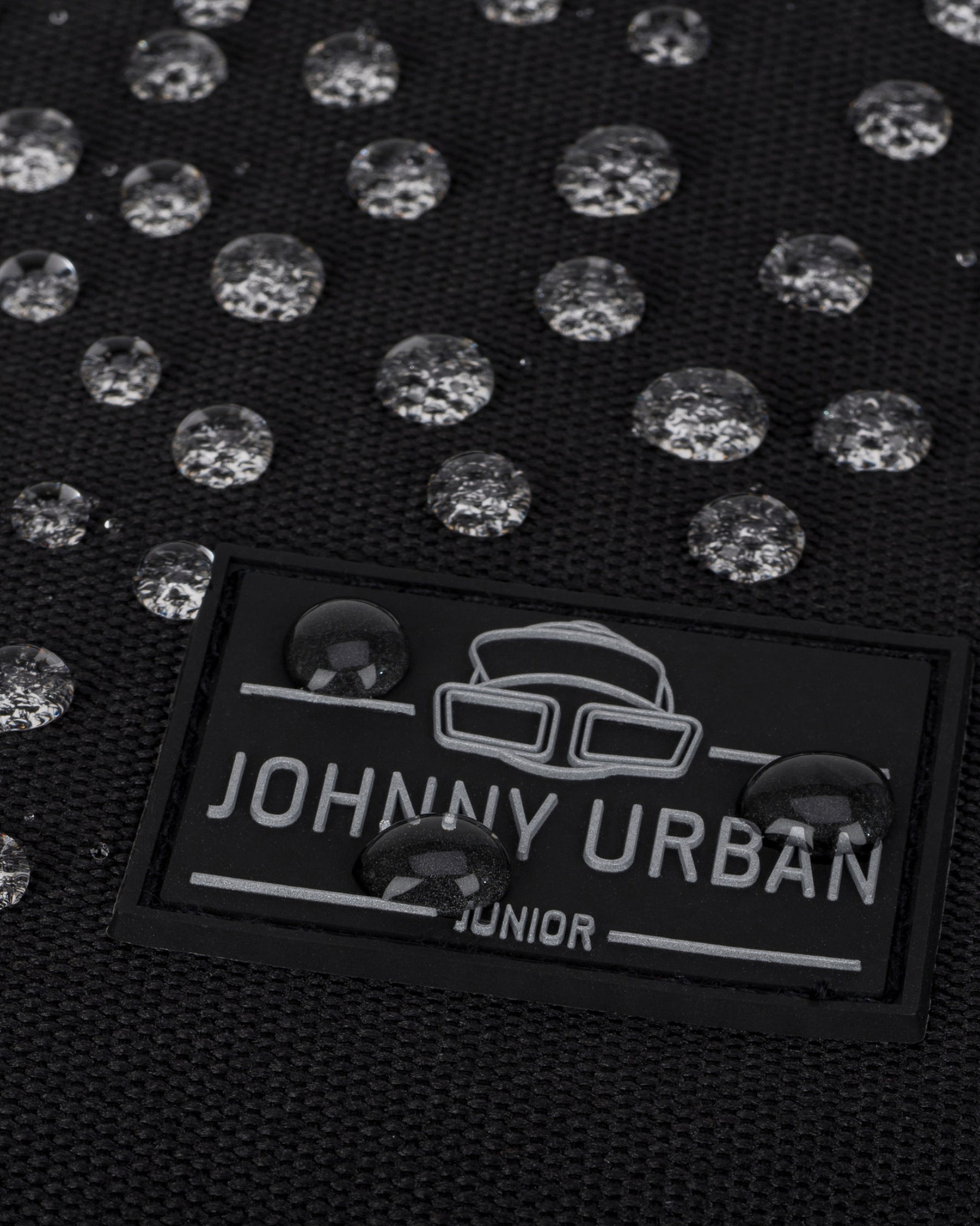 Nahaufnahme eines schwarzen wasserdichten Rolltop Kinderrucksacks 'Aaron' mit Johnny Urban Junior Logo und Wasserperlen