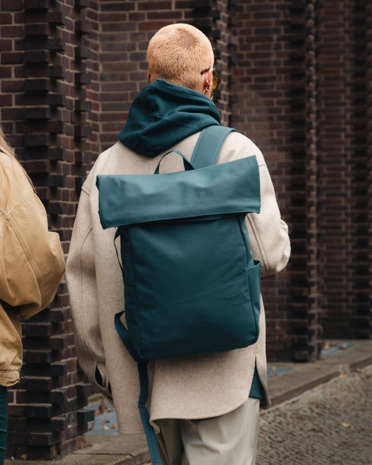 Frau mit kurzem blondem Haar trägt petrolfarbenen Rolltop Rucksack 'Henry' vor Ziegelmauer