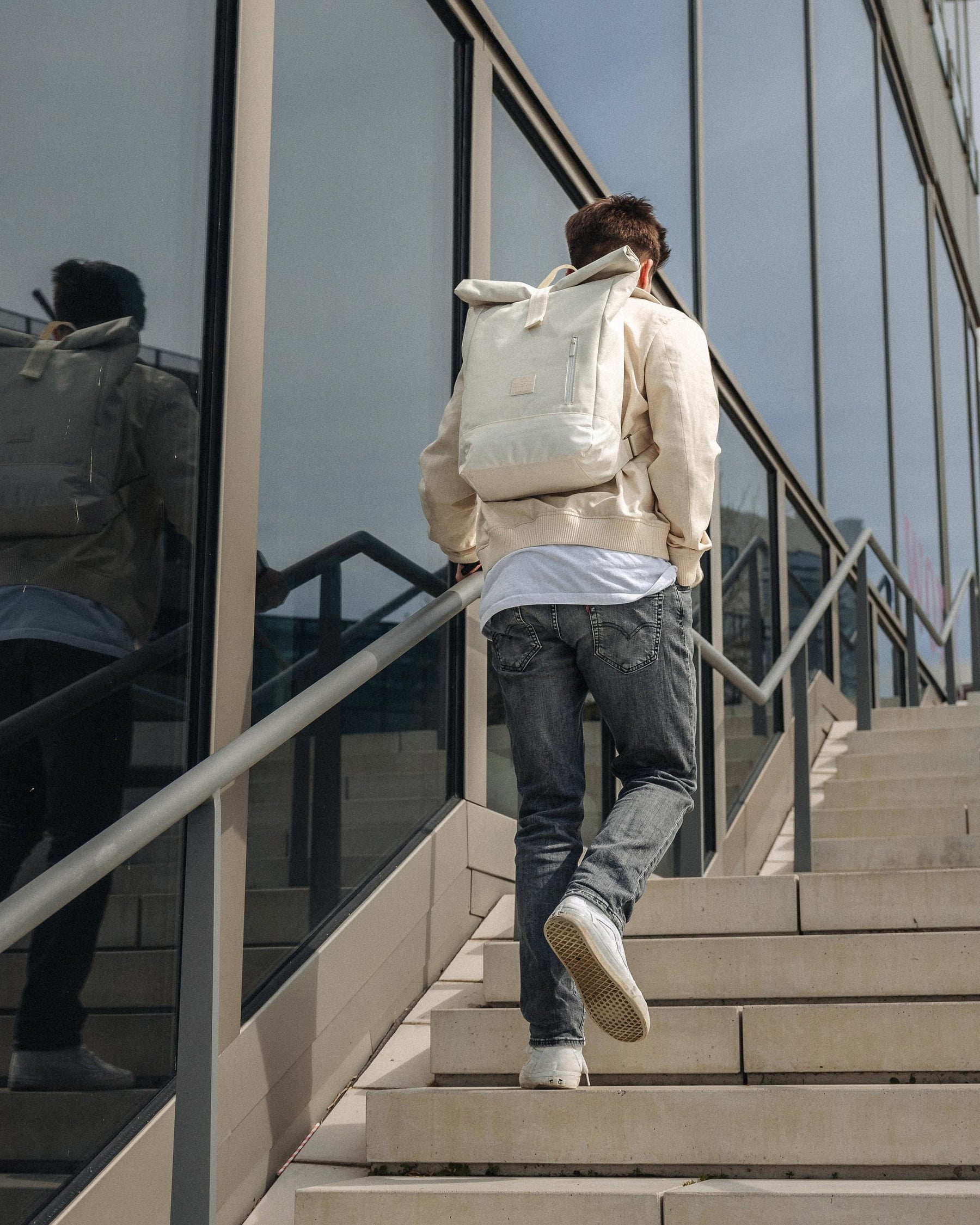 Mann trägt weißen Rolltop-Rucksack 'Robin Medium' und steigt Außenstiege neben Glasfassade hoch