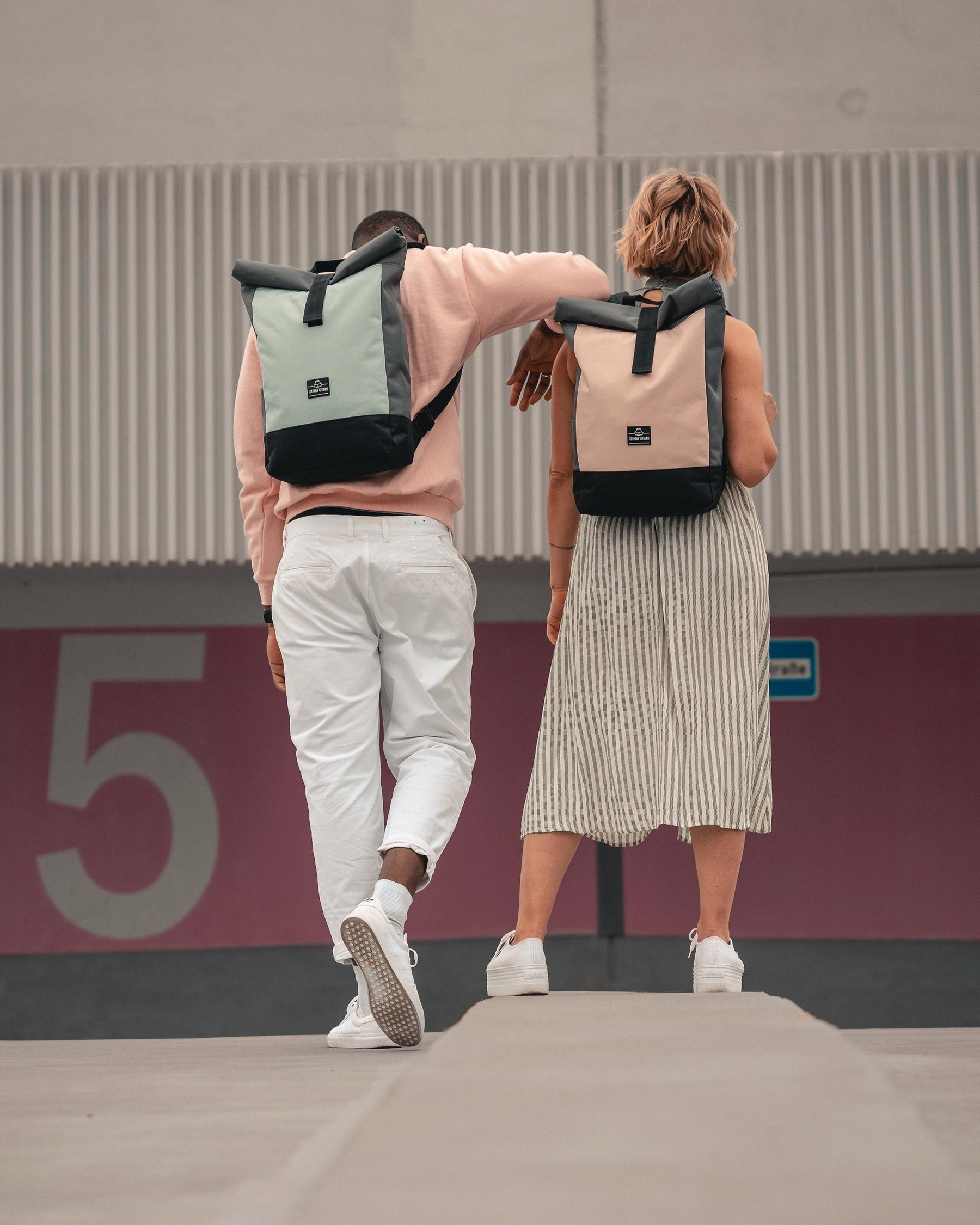 Zwei Personen von hinten mit Rolltop Rucksack 'Ryan' in Pastellfarben, stehend vor einer Wand mit großer Zahl 5