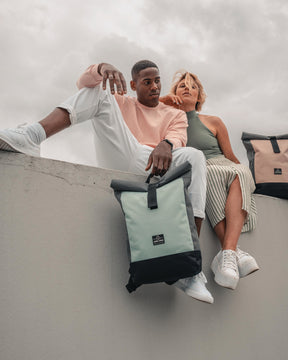 Junger Mann und Frau sitzen auf einer Mauer mit Rolltop Rucksäcken 'Ryan' in Pastell-Mint und Pastell-Rosa