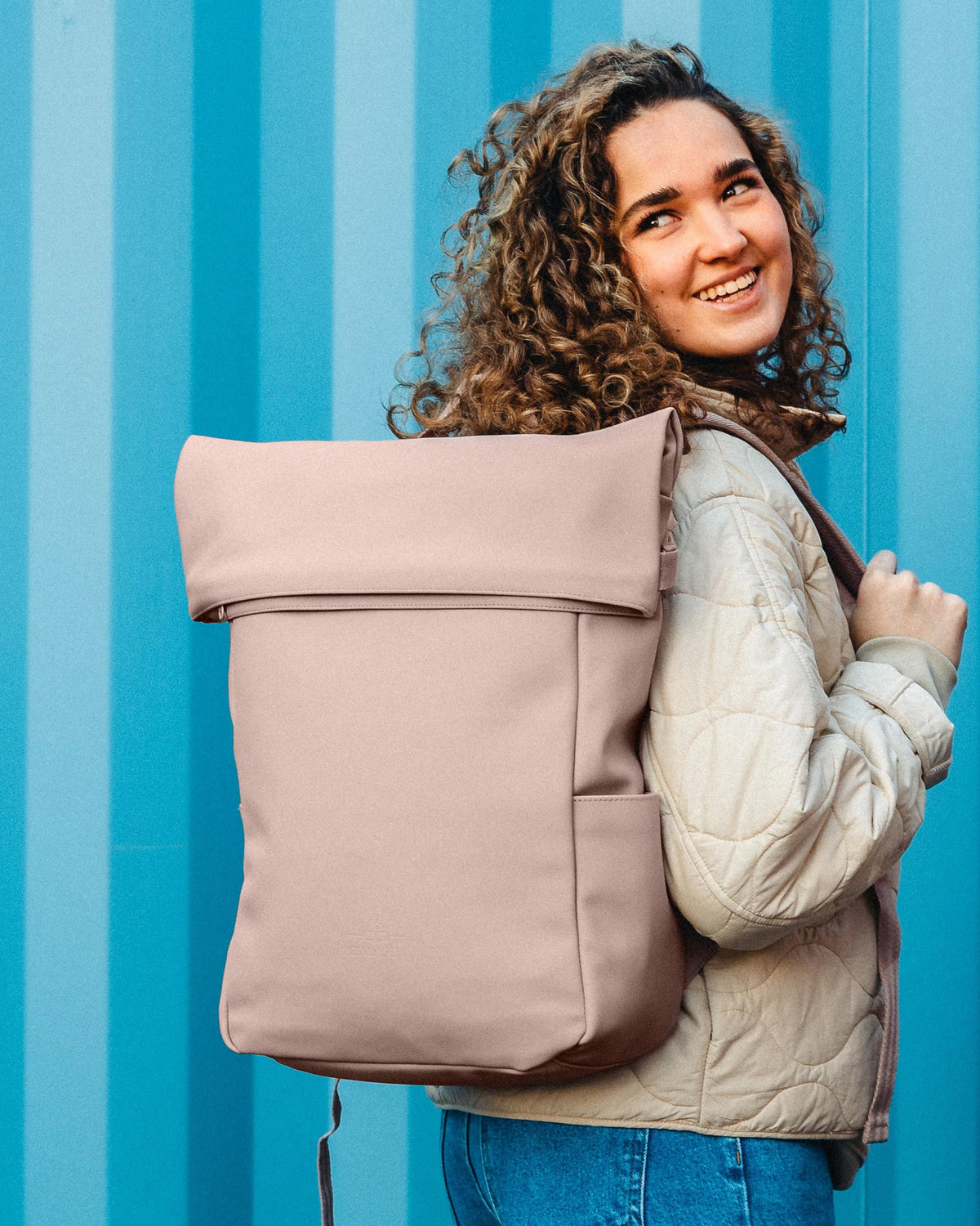 Frau mit lockigem Haar trägt rosa Johnny Urban Rolltop Rucksack Henry vor blau gestreiftem Hintergrund