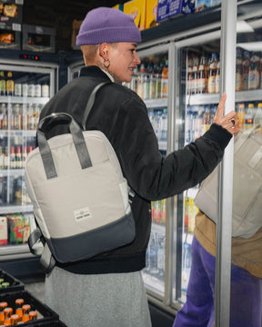 Frau mit lila Mütze und schwarzer Jacke trägt den beigen-grauen Rucksack 'Jona Medium' von Johnny Urban beim Öffnen einer Kühlschranktür