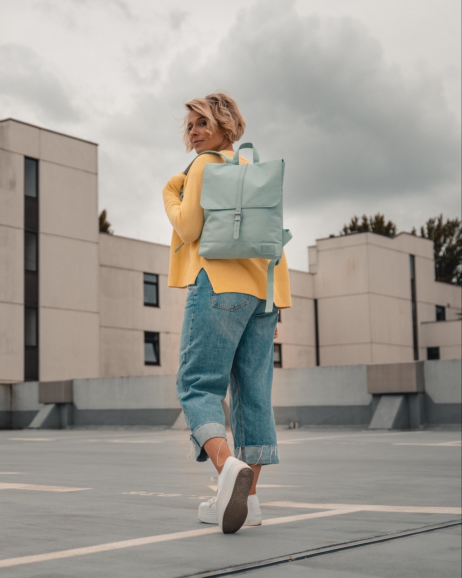 Frau trägt mintfarbigen Rucksack 'Mia', gelber Pullover, blaue Jeans, weiße Sneakers auf Dachparkplatz