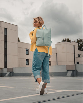 Frau trägt mintfarbigen Rucksack 'Mia', gelber Pullover, blaue Jeans, weiße Sneakers auf Dachparkplatz