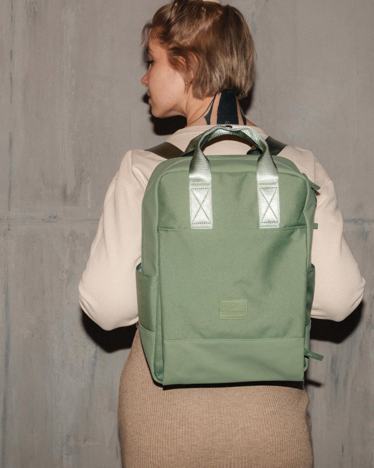 Frau trägt grünen nachhaltigen Rucksack 'Jona Large' von Johnny Urban vor grauer Wand