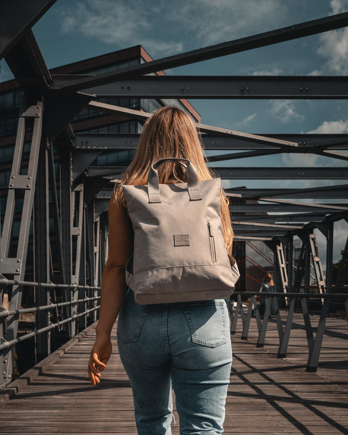 Frau trägt beige Rucksacktasche 'Ivy' von Johnny Urban auf Holzbrücke im urbanen Umfeld
