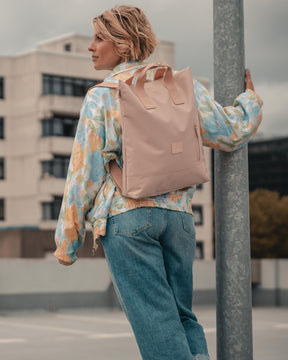 Frau trägt rosa Rucksacktasche 'Ivy' von Johnny Urban, lässige bunte Jacke und blaue Jeans, stützt sich auf Laternenpfahl