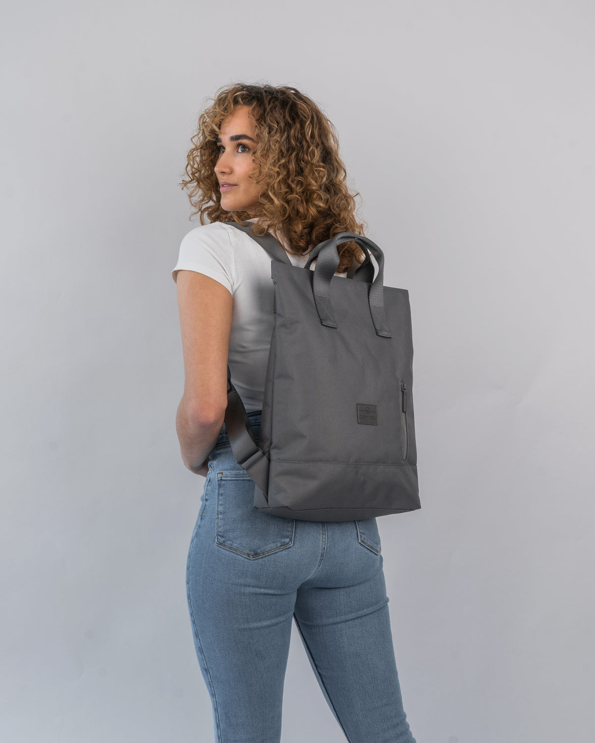 Frau mit lockigem Haar trägt dunkelgrauen Rucksacktasche 'Ivy' von Johnny Urban über weißen T-Shirt und Jeans