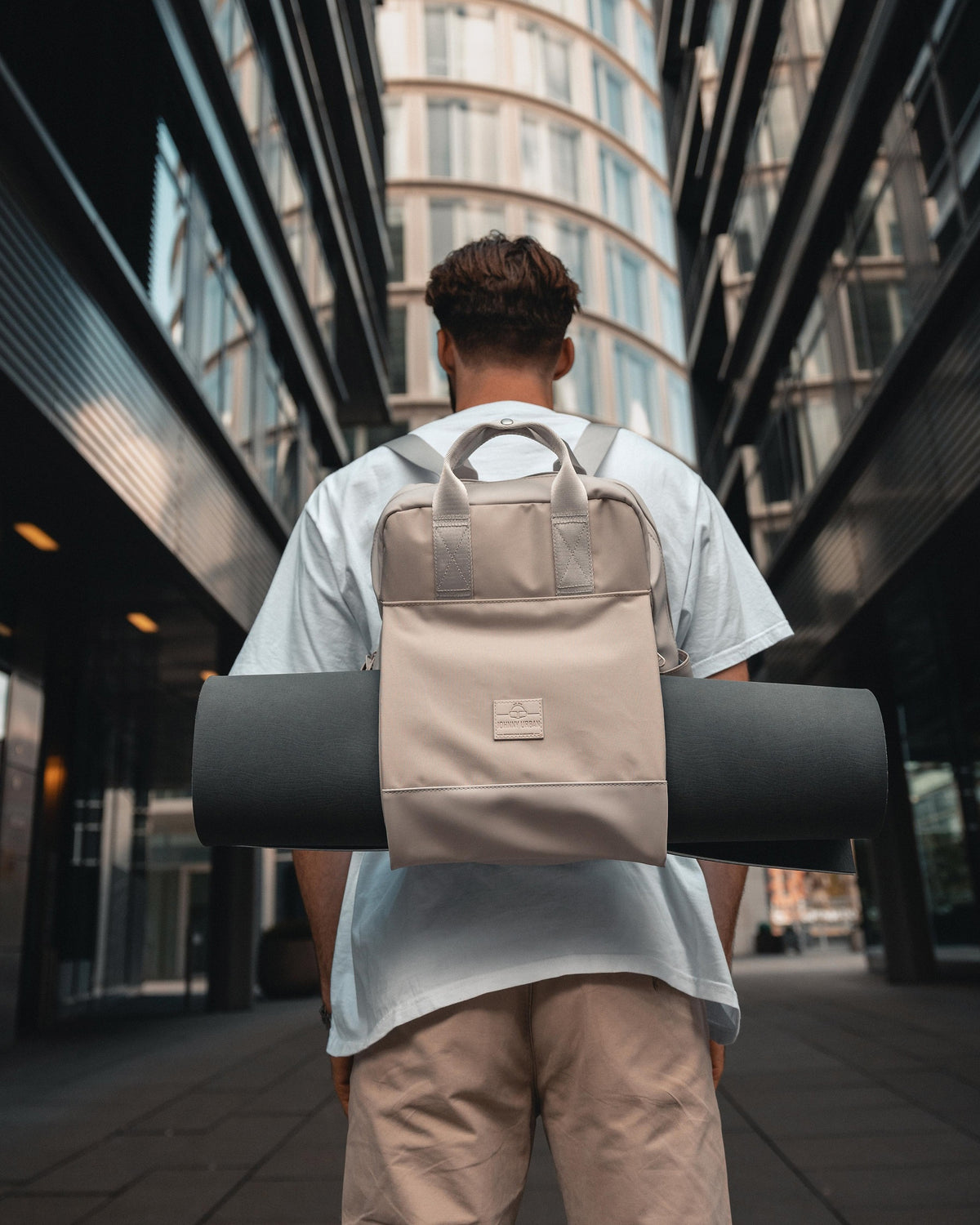 Mann mit beigem Rucksack 'Jona Large' trägt schwarze Yogamatte in urbaner Umgebung