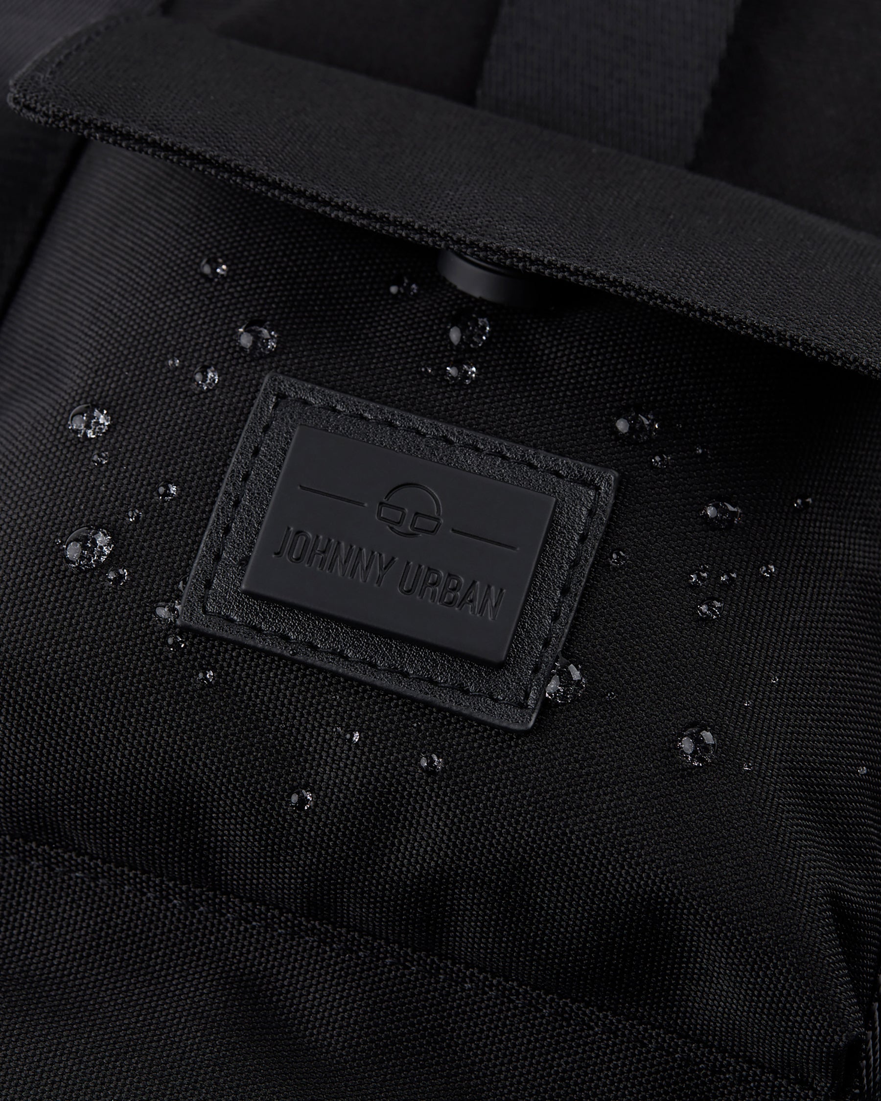 Nahaufnahme des schwarzen wasserabweisenden Rucksacks 'Jules' mit Johnny Urban Logo und Wassertröpfchen auf dem Stoff