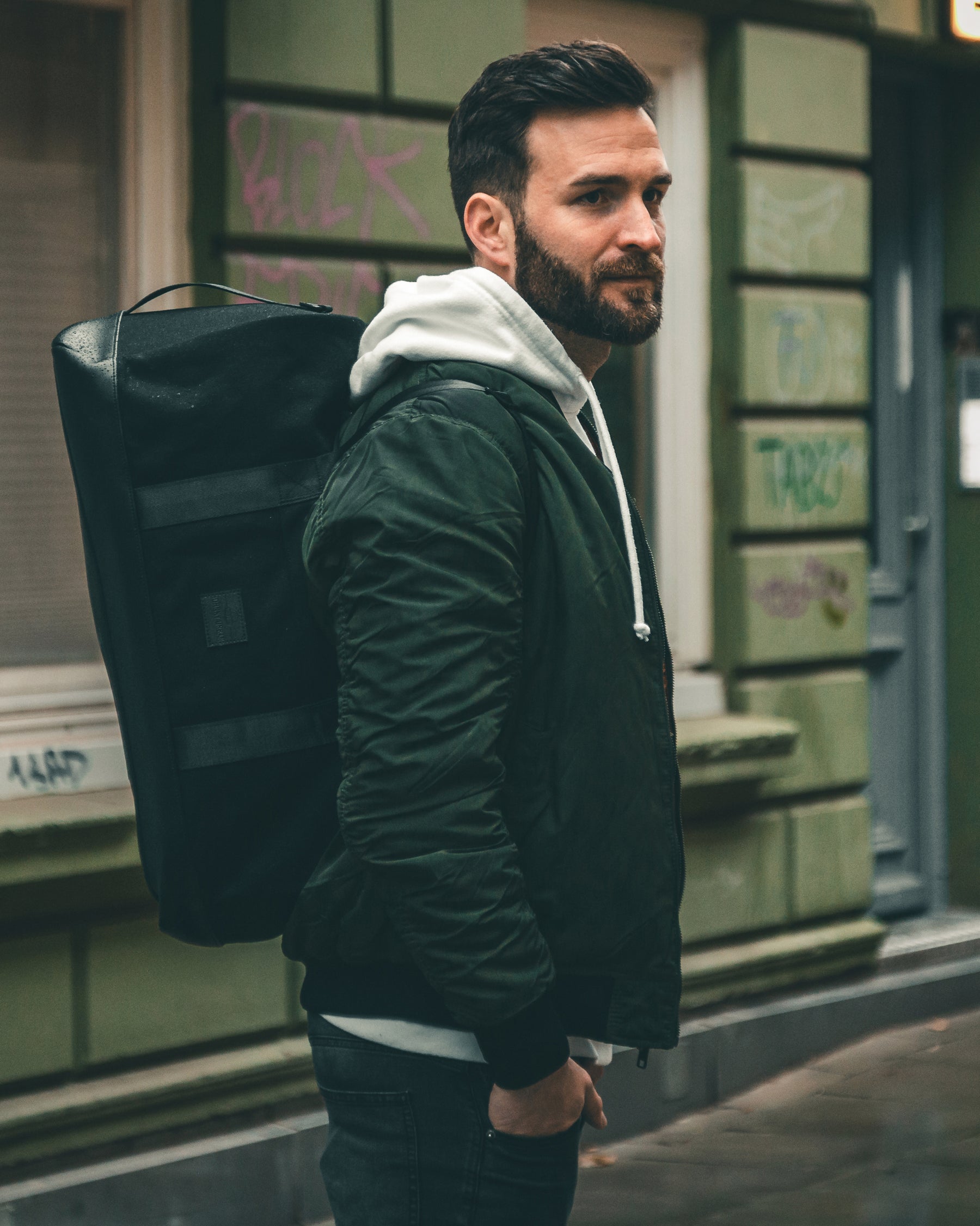 Mann mit schwarzem Weekender-Rucksack 'Paul' von Johnny Urban, grüne Jacke und grauer Hoodie in urbaner Umgebung